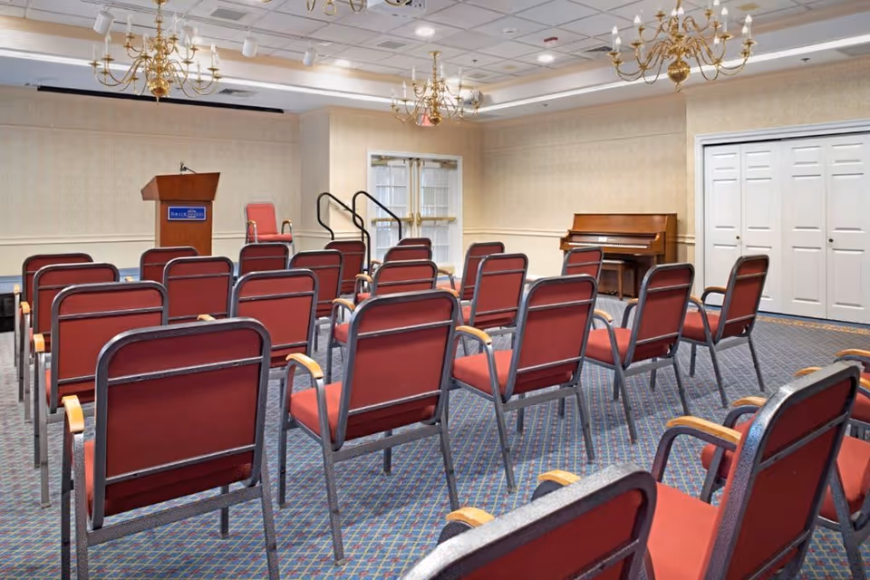 A small conference or meeting room with rows of red cushioned chairs facing a wooden podium. The room has beige wallpaper, a blue patterned carpet, two chandeliers, a piano against the wall, and double glass doors at the front.