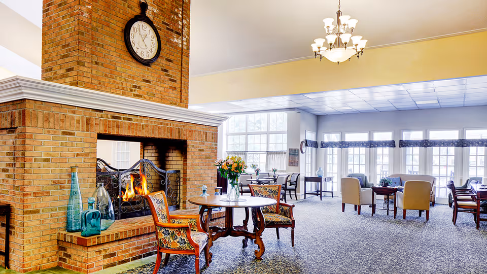 A spacious and well-lit common area with a large brick fireplace in the center, featuring a clock above it. The room has multiple seating arrangements including floral upholstered chairs around a wooden table, beige armchairs around a coffee table, and dining tables with chairs near large windows letting in natural light.