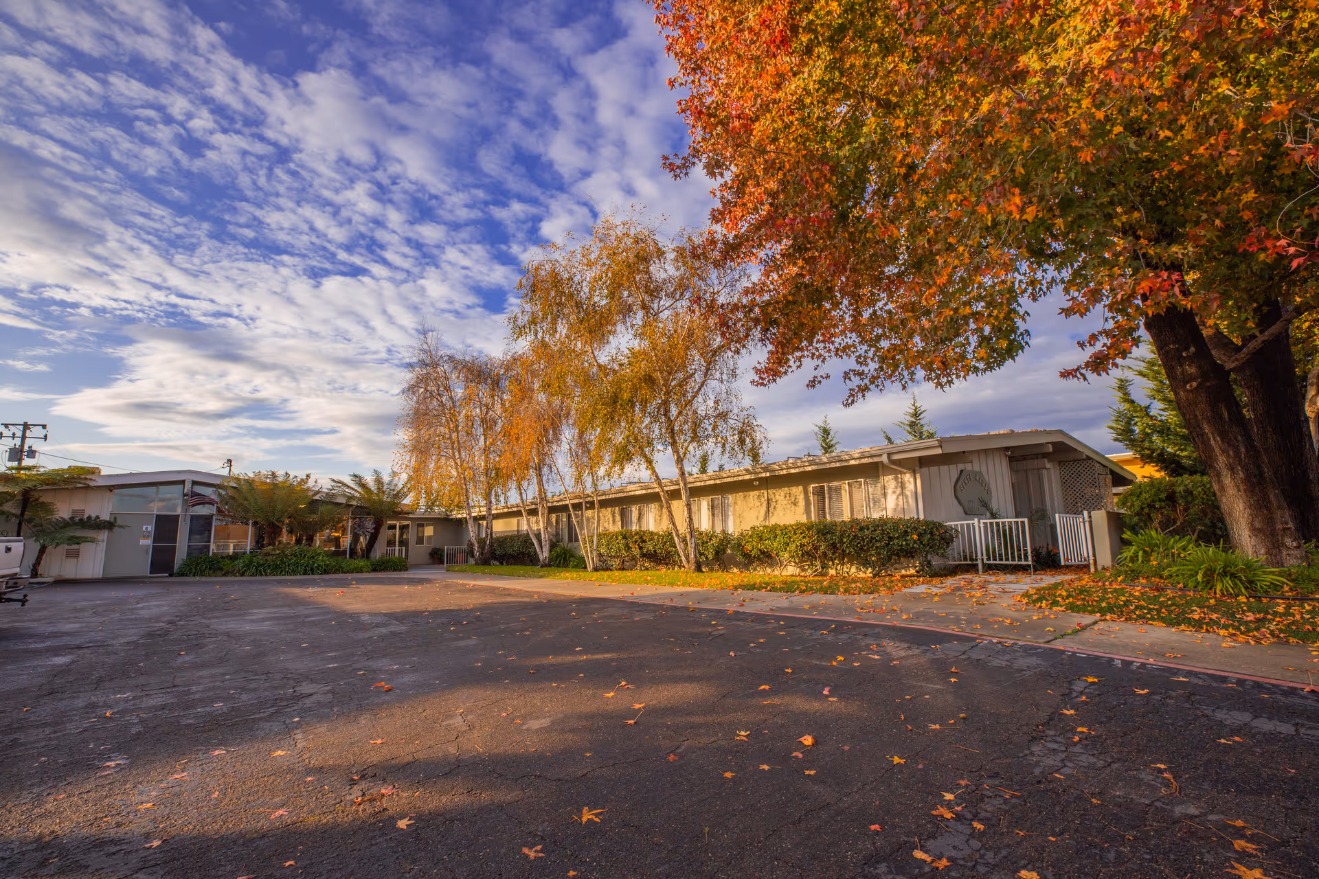Single-story senior living facility building with autumn trees, a paved driveway, and a blue sky.