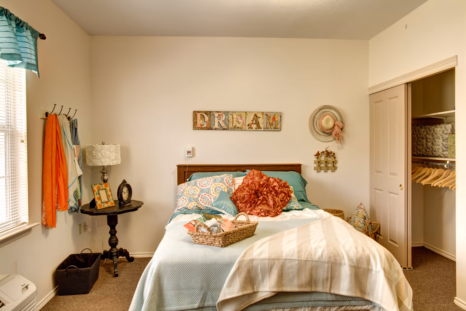 A cozy bedroom with a neatly made bed featuring decorative pillows and a basket with magazines on top. The wall above the bed has a colorful sign that reads 'DREAM' and a hat hanging on a hook. To the left, there is a small round table with a lamp, a picture frame, and a clock. A window with teal curtains lets in natural light. On the right side, there is an open closet with hangers and storage boxes.