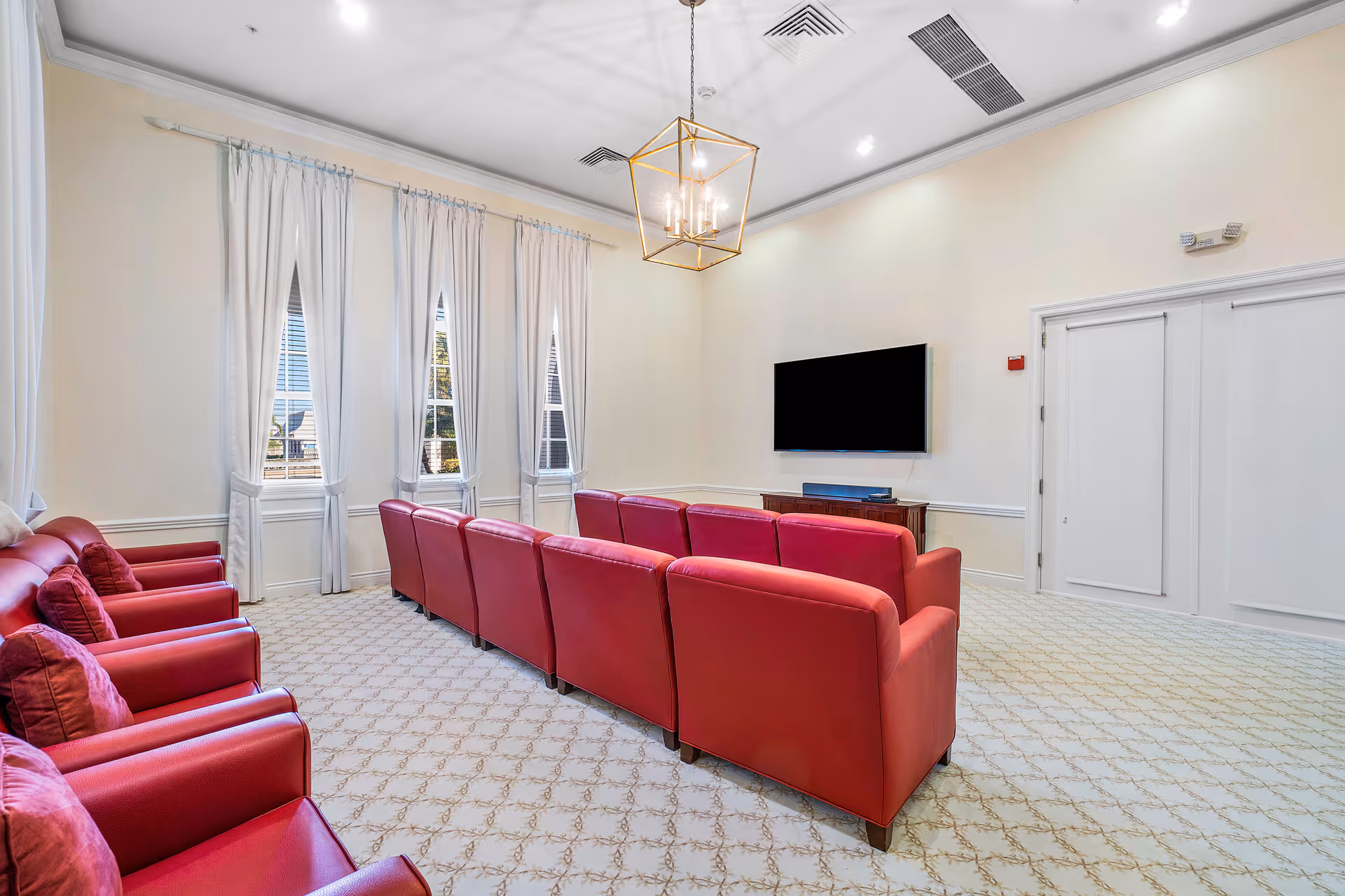 A bright and spacious common room with cream-colored walls and carpet, featuring two rows of red upholstered armchairs facing a wall-mounted flat-screen TV. The room has three tall windows with white curtains, a modern gold chandelier hanging from the ceiling, and a wooden console table beneath the TV.