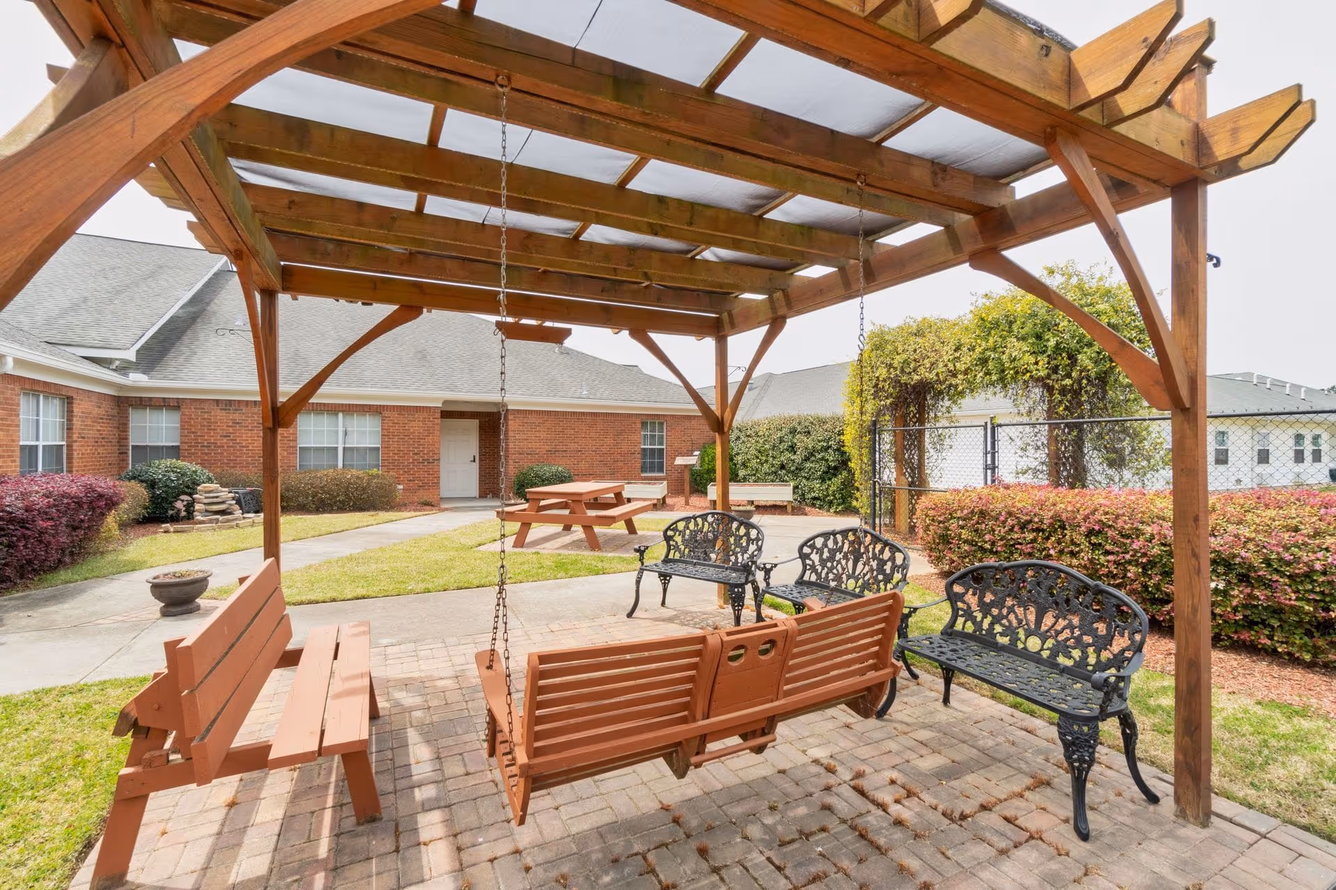 Covered wooden pergola with a hanging swing, metal benches, and a picnic table in a landscaped courtyard in front of a brick building.