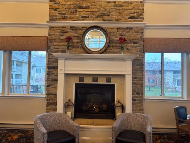 A cozy interior space featuring a stone fireplace with a white mantel. Above the mantel is a round mirror flanked by two vases each holding a red flower. Two upholstered chairs are positioned in front of the fireplace. Large windows on either side of the fireplace show an outdoor view of residential buildings and greenery.