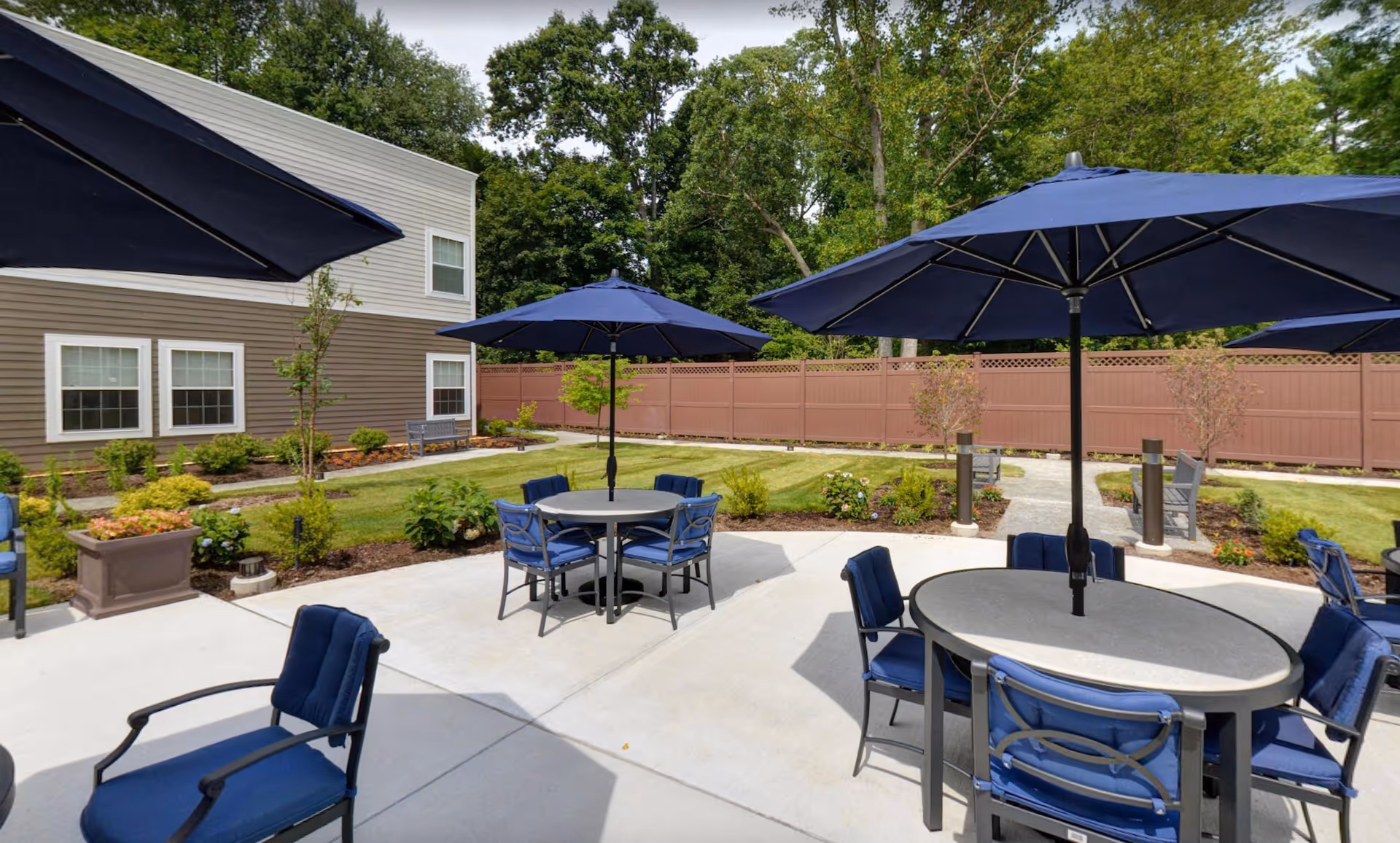 Outdoor patio area at a senior living facility with round tables, blue cushioned chairs, and large blue umbrellas. The patio is surrounded by a garden with small trees and shrubs, a brown privacy fence, and a two-story building with beige and brown siding in the background.