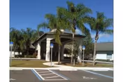 Entrance area of Sunny Hills Assisted Living Facility Homestead with a covered drop-off zone, palm trees, and designated handicap parking spaces under a clear blue sky.