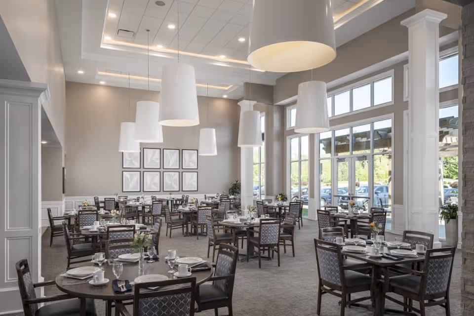 A spacious dining room with multiple round tables set with plates, cups, and silverware. The room features large windows allowing natural light to fill the space, modern hanging light fixtures, and framed artwork on the far wall. The chairs have patterned upholstery and the overall decor is elegant and inviting.