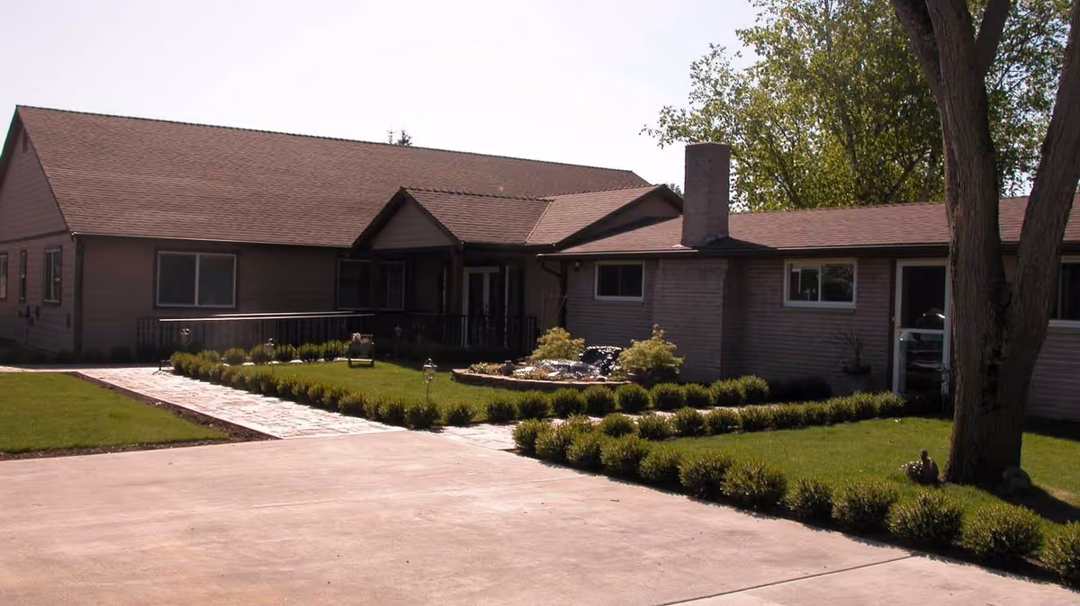 Single-story building with a brown roof and beige walls, surrounded by a well-maintained lawn and shrubbery. A paved walkway leads to the entrance, and there is a large tree on the right side of the image.