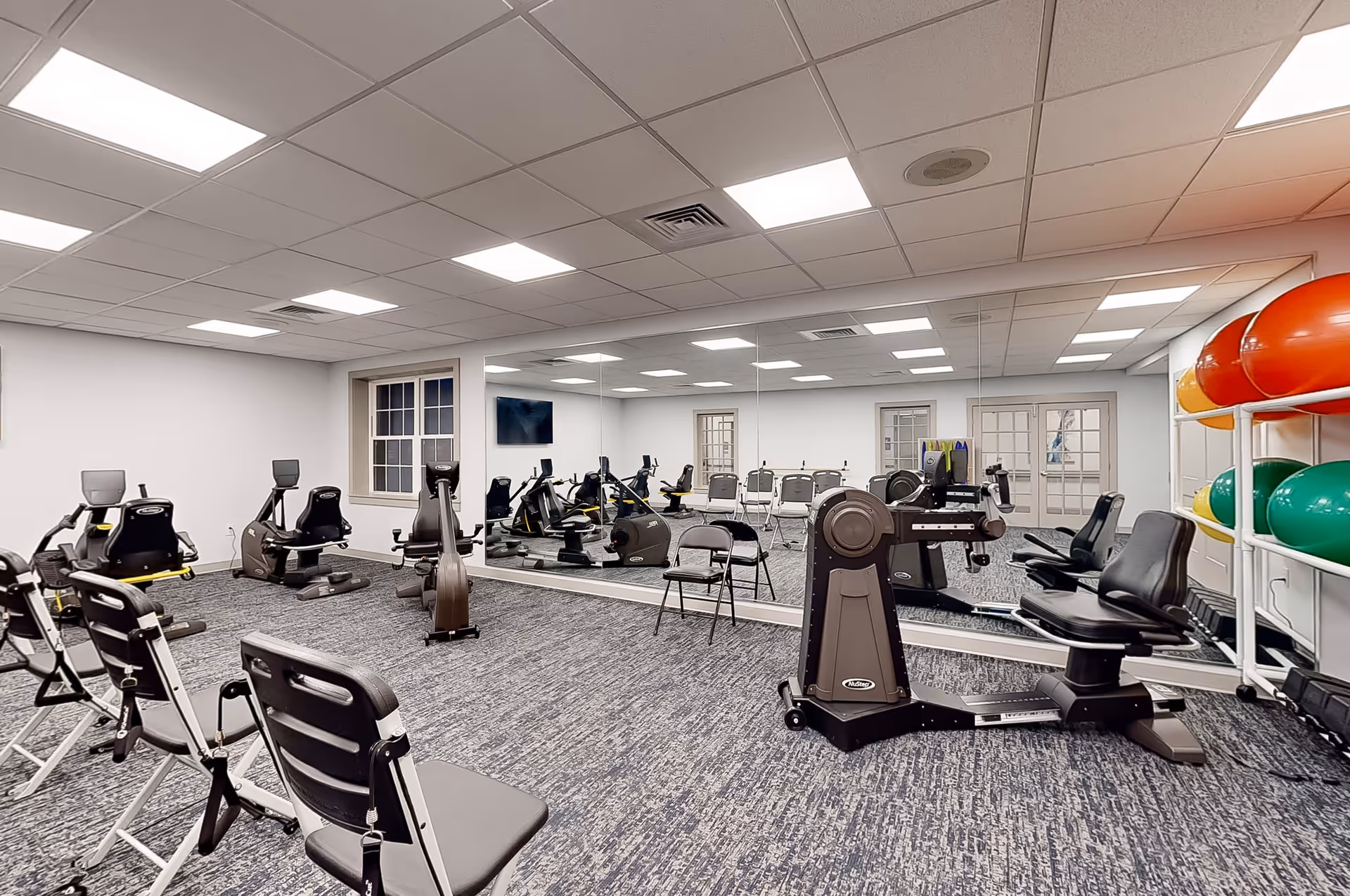 A fitness room with exercise equipment including stationary bikes and rowing machines, several chairs, a large wall mirror, and a rack holding colorful exercise balls. The room has a carpeted floor, white walls, and a ceiling with recessed lighting.