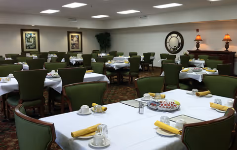 A dining room with multiple tables covered in white tablecloths, each set with cups, glasses, yellow napkins, and silverware. The chairs are green with wooden frames. The room has framed artwork on the walls, a large round mirror, two lamps on a wooden sideboard, and a potted plant in the corner.
