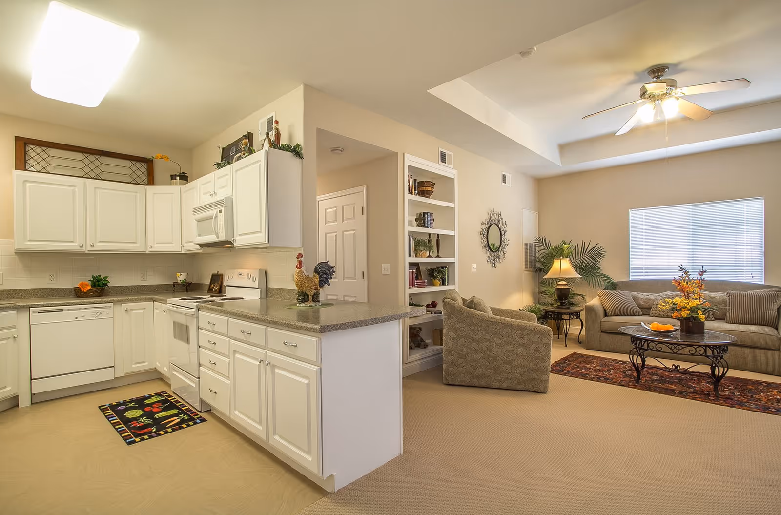 A bright and spacious open-concept living area and kitchen in a senior living community. The kitchen features white cabinets, a dishwasher, stove, microwave, and a countertop with decorative items including a rooster figurine. The living room has a beige sofa, an armchair, a coffee table with a floral arrangement, a side table with a lamp, a ceiling fan, and a window with blinds. There is a built-in bookshelf with various decor items and a decorative wall mirror.
