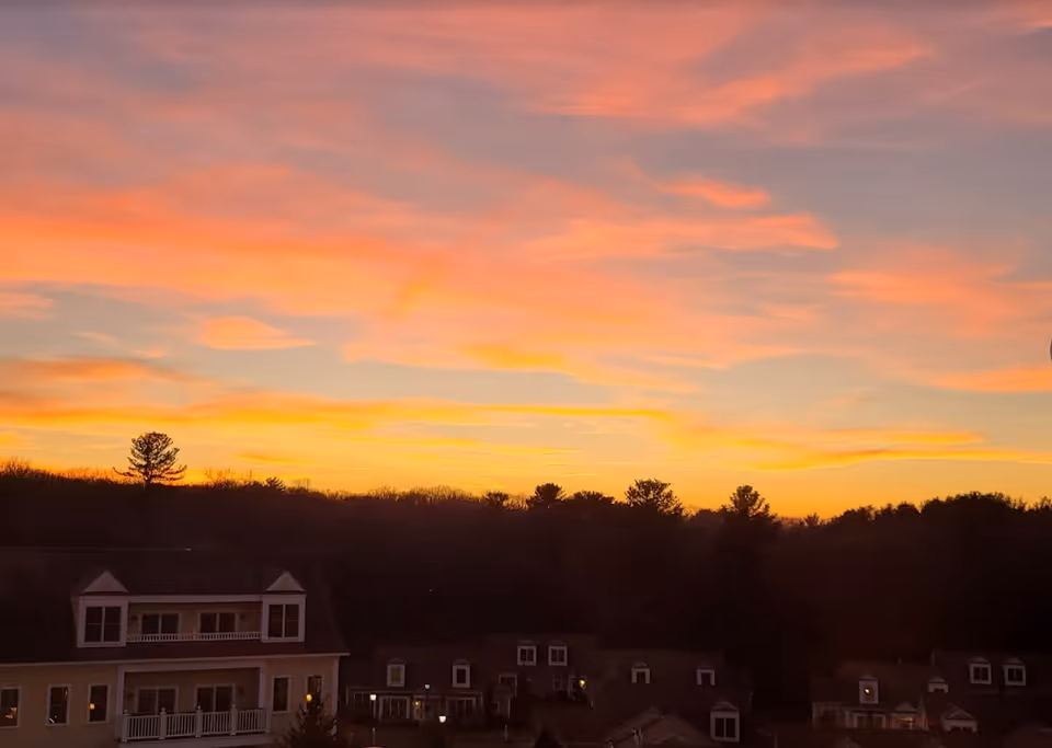 Sunset sky with vibrant orange and pink clouds above a silhouette of trees and a residential building with multiple windows and balconies.