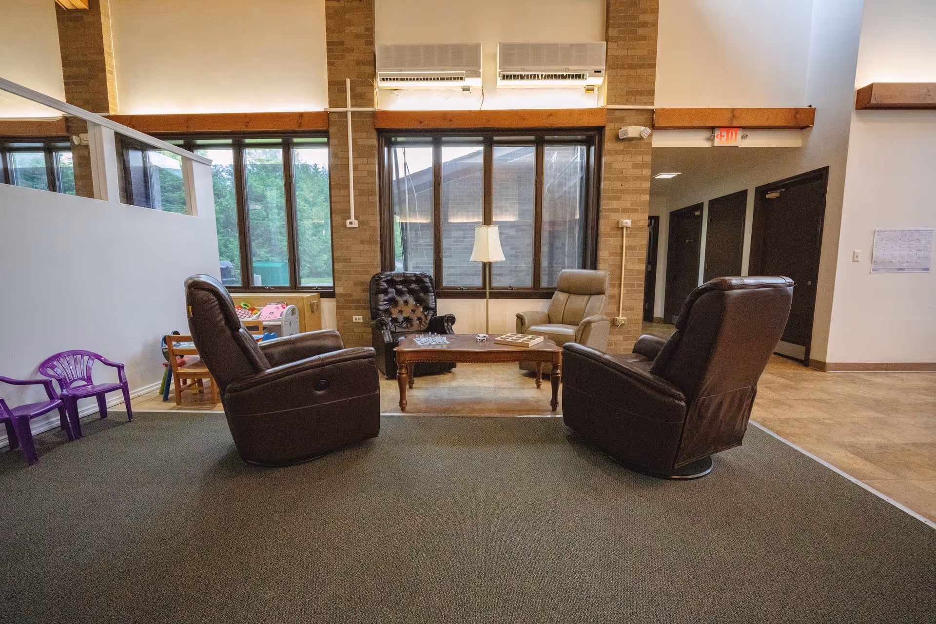 A cozy seating area in a senior living facility with four armchairs arranged around a wooden coffee table. The table has a chess set and a board game on it. Large windows with views of greenery outside and brick walls surround the space. There is a floor lamp between two of the chairs and a small play area with children's furniture and toys to the left.