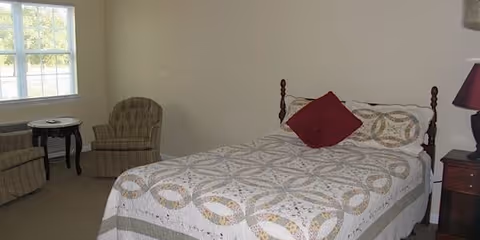 A bedroom with a bed covered in a patterned quilt with a red pillow, two upholstered chairs near a window, a small round table between the chairs, and a wooden nightstand with a lamp.