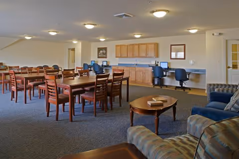 A spacious room with multiple wooden tables and chairs arranged for group seating. Along one wall, there are three computer workstations with office chairs and wooden cabinets above a countertop. In the foreground, there are cushioned armchairs and a wooden coffee table with books on it. The room has carpeted flooring and ceiling lights.