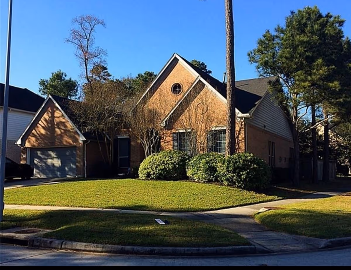 Exterior view of a single-story brick house with a garage, surrounded by green bushes and trees under a clear blue sky.