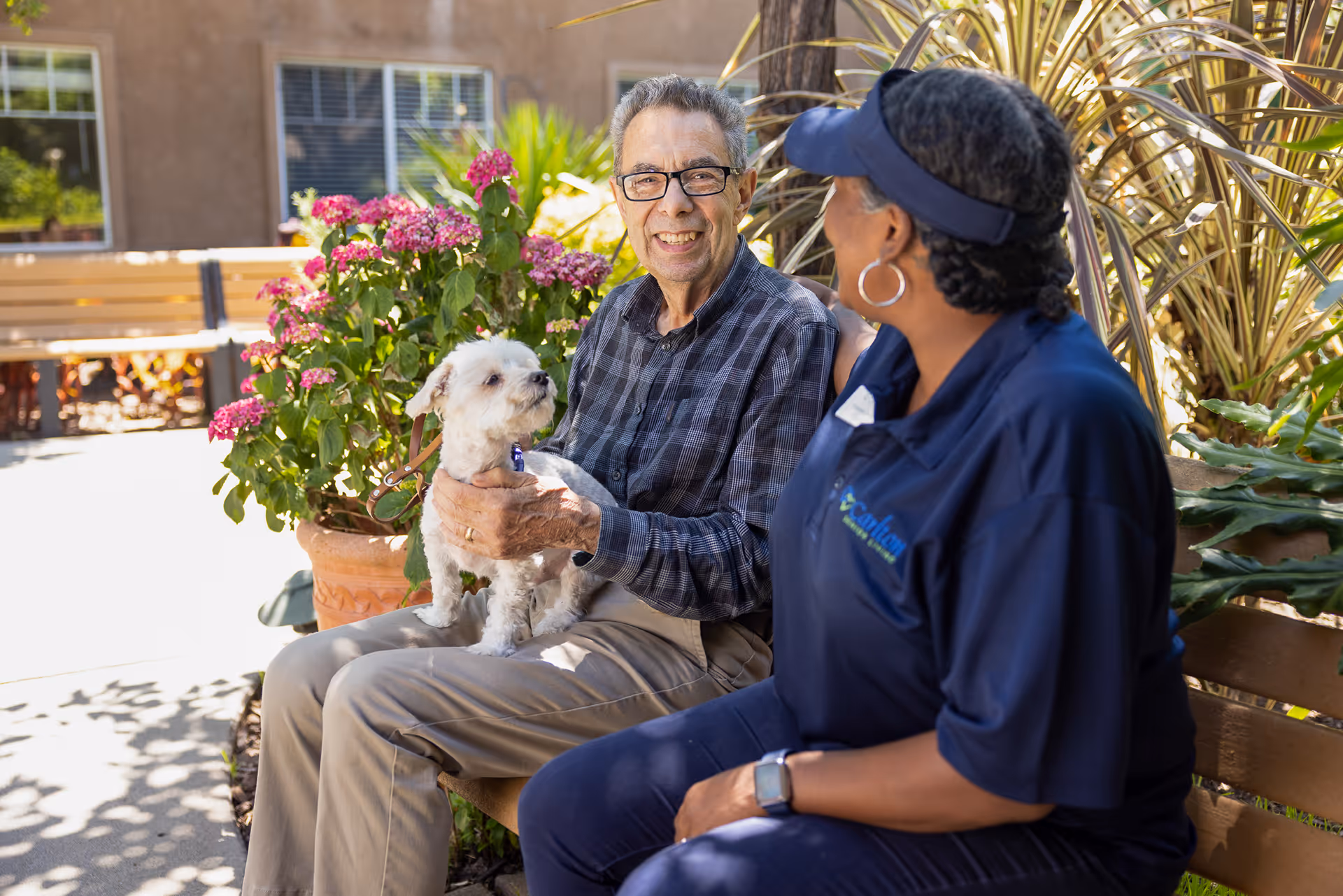 An elderly man sitting on a bench outdoors holding a small white dog, smiling and looking at a woman in a blue uniform sitting next to him. They are surrounded by plants and flowers in a sunny garden area.
