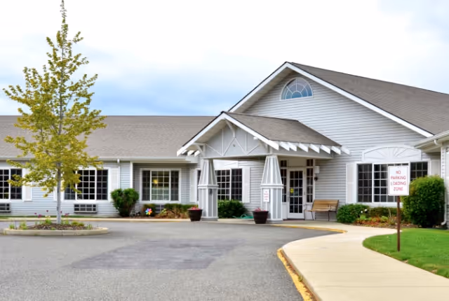 Exterior view of a single-story senior living facility building with white siding, a peaked roof, and a covered entrance supported by columns. There is a small tree and some shrubs in front, a paved driveway, a sidewalk, and a sign indicating no parking in the loading zone.