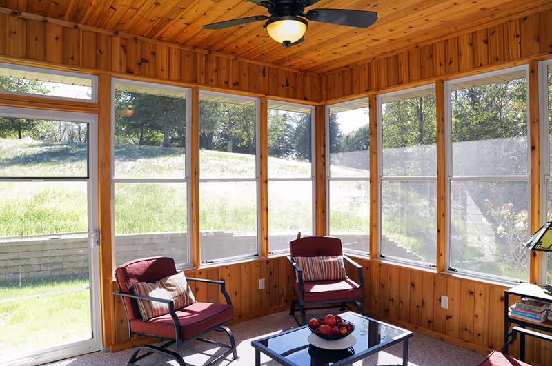 Sunroom with wood-paneled walls and ceiling, large windows, two red cushioned chairs and a coffee table with a bowl of apples.