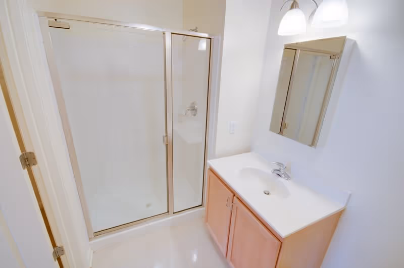 A clean bathroom featuring a glass-enclosed shower with sliding doors, a white countertop with an integrated sink, wooden cabinetry below, a wall-mounted mirror, and a two-light fixture above the mirror.