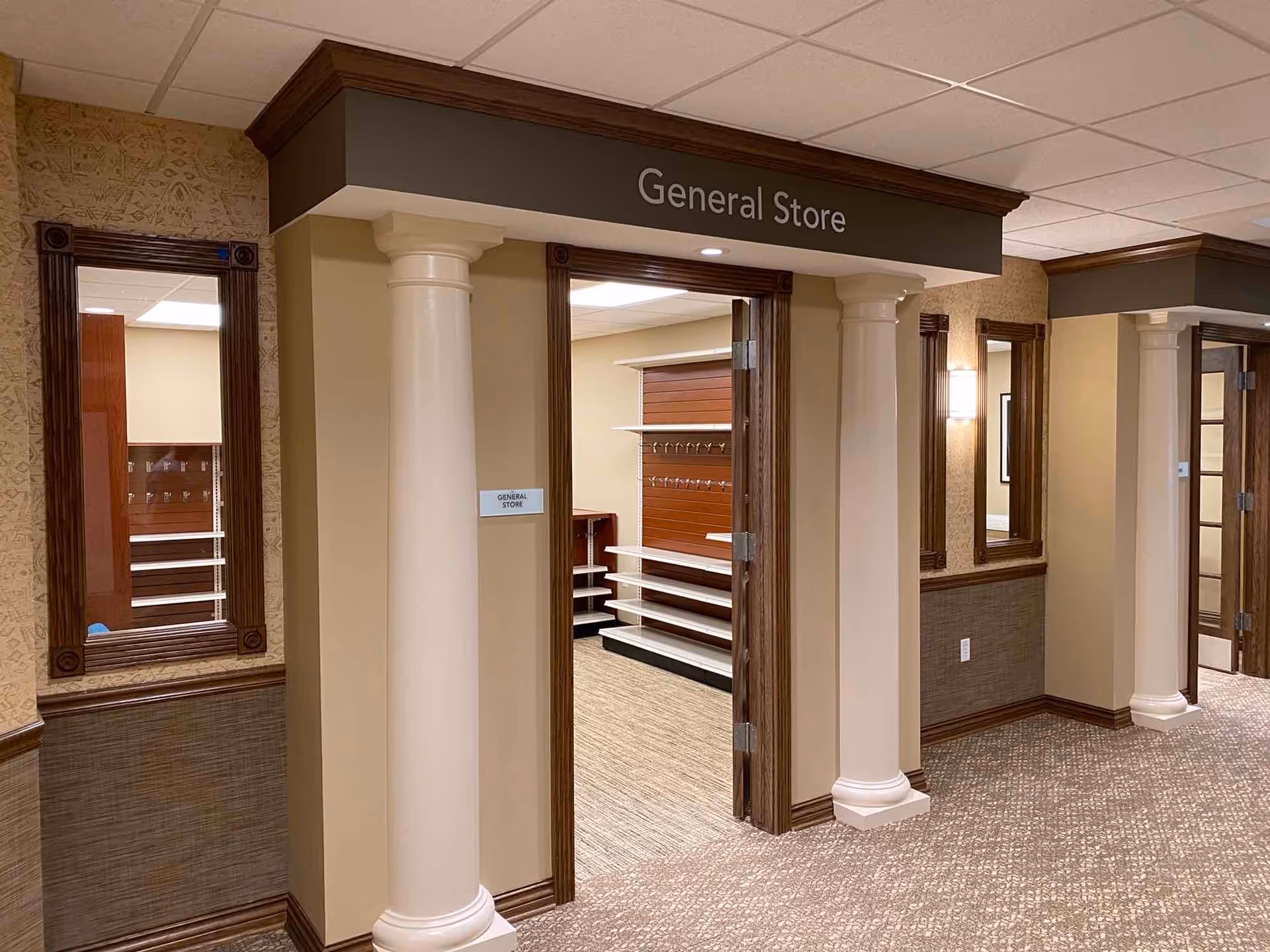 Interior view of a hallway in a senior living facility with a doorway labeled 'General Store'. The entrance is framed by two white columns and dark wood trim. Inside, empty shelves and display racks are visible. The hallway has carpeted flooring, beige walls with decorative wainscoting, and mirrors on the walls.