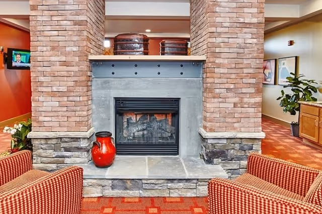 Communal lounge area with a central double-sided fireplace set between brick columns, red patterned chairs, and decorative accents.