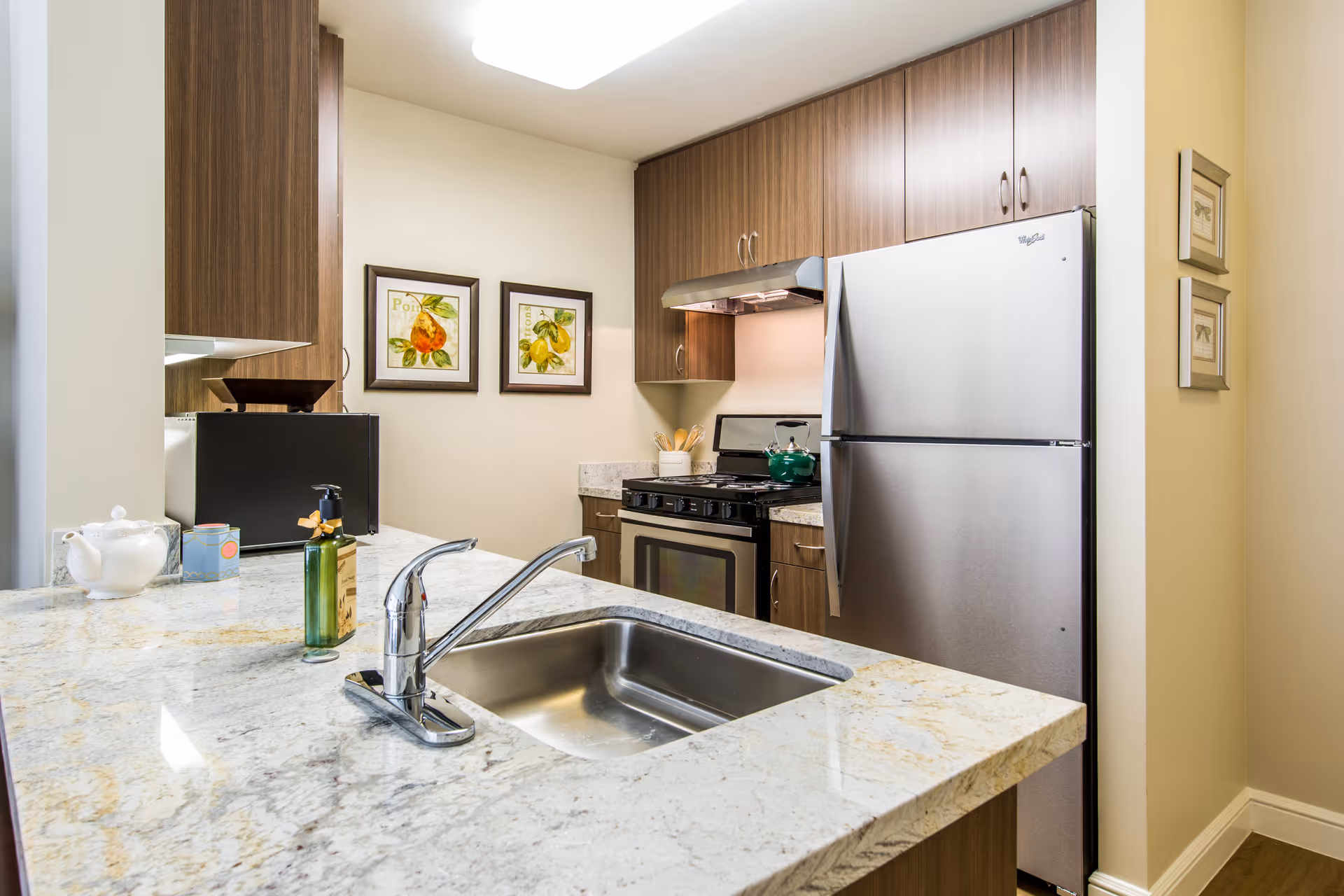 Modern kitchen with granite countertops, a stainless steel sink and faucet, a stainless steel refrigerator, and a stove with a green kettle on it. The kitchen has wooden cabinets and two framed fruit pictures on the wall.