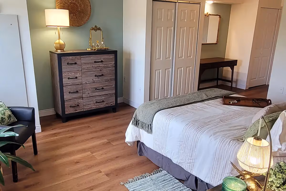 A cozy bedroom featuring a bed with white and green bedding, a wooden dresser with a lamp and decorative items on top, a small desk with a mirror above it, a black chair with a green cushion, and wooden flooring.