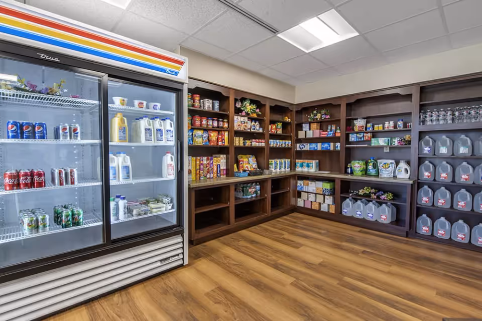 Interior view of a small convenience store or pantry area with wooden shelves stocked with various food items, beverages, and household supplies. A glass-door refrigerator on the left contains milk, soda cans, and other drinks. The floor is wooden, and the ceiling has white tiles with fluorescent lighting.