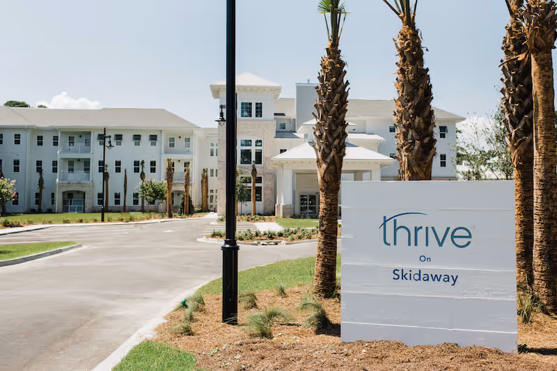 Front exterior of the Thrive on Skidaway senior living building with entrance, palm trees, and a sign.