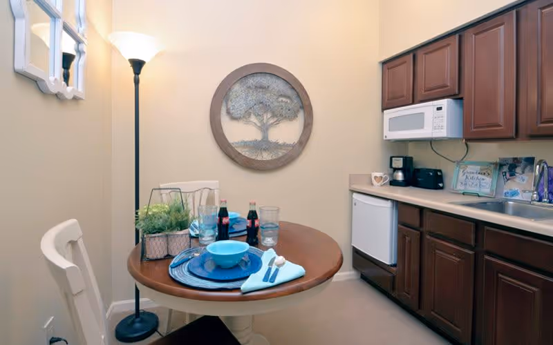 A small kitchen and dining area with a round wooden table set for one with blue dishware, utensils, and two bottles of soda. The kitchen has dark wooden cabinets, a white microwave, a coffee maker, a toaster, and a small refrigerator. A floor lamp and a decorative wall art featuring a tree are also visible.
