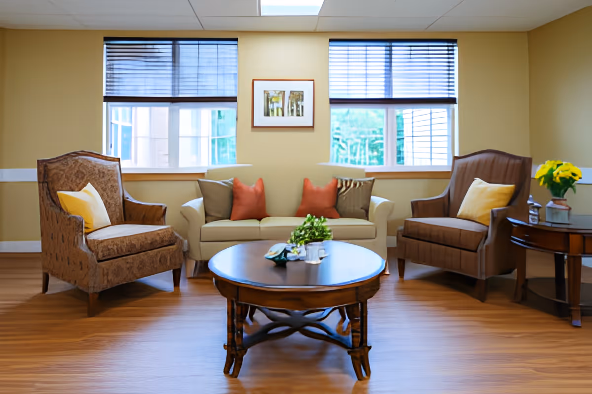 A cozy living room with a beige sofa adorned with four cushions in red, gray, and striped patterns. On either side of the sofa are two brown armchairs, each with a yellow pillow. In front of the seating area is a round wooden coffee table with a small plant and decorative items on it. Behind the sofa are two windows with blinds partially drawn, and a framed picture hangs on the wall between the windows. To the right, there is a side table with a vase of yellow flowers. The room has wooden flooring and light yellow walls.