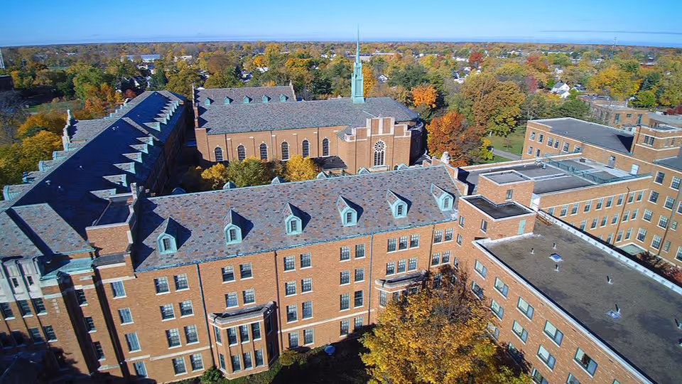 Aerial view of a large senior living facility complex with multiple connected brick buildings, surrounded by trees with autumn foliage under a clear blue sky.