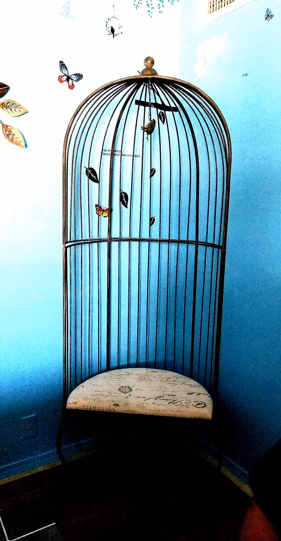 A decorative birdcage-shaped chair with a cushioned seat featuring script writing, placed in a corner against light blue walls adorned with butterfly and leaf wall decals.