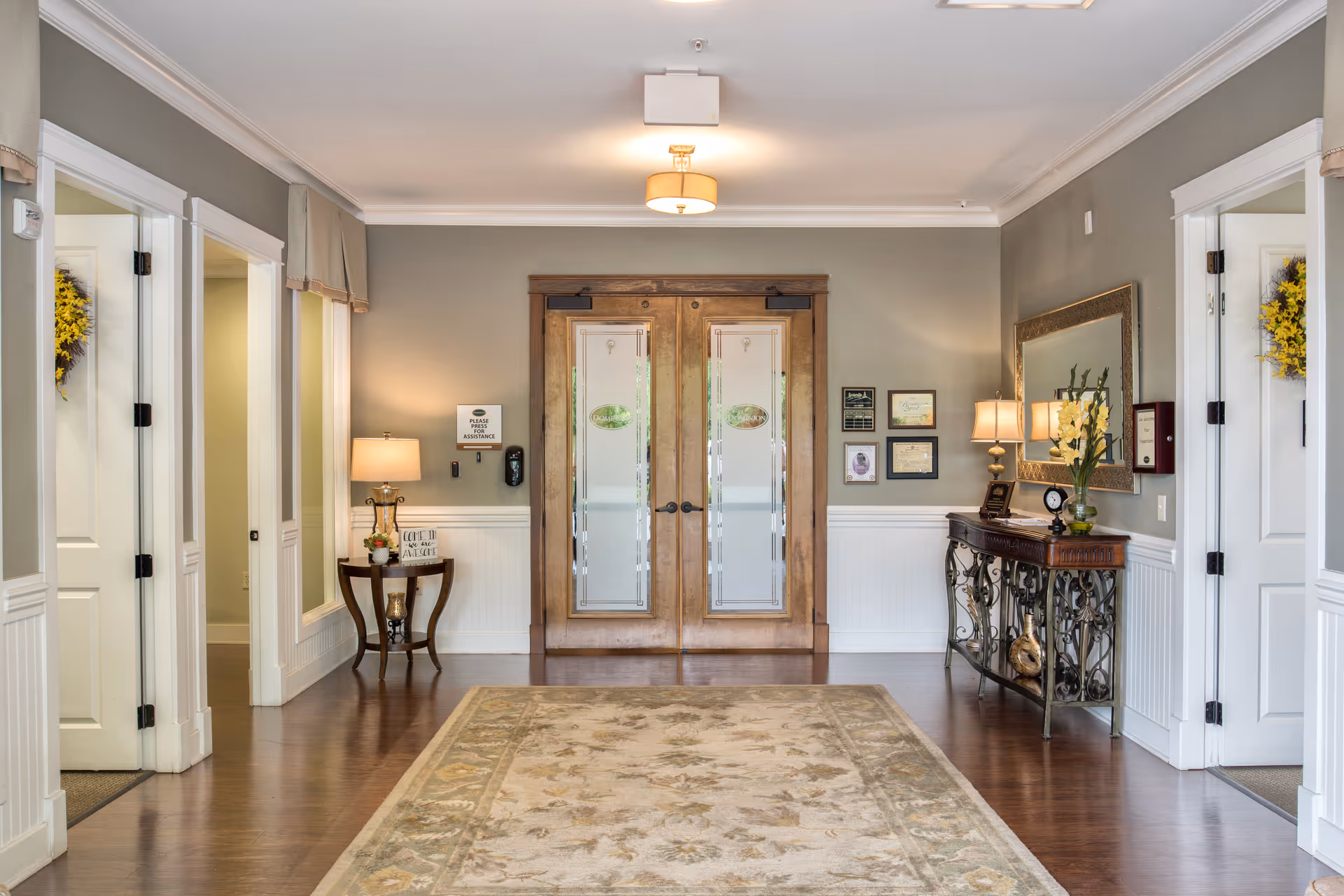 A well-lit interior hallway with wooden double doors at the end, flanked by side rooms with white doors. The hallway features a large patterned rug on a wooden floor, a small table with a lamp and decorative items on the left, and a console table with a lamp, flowers, and framed certificates on the right. The walls are painted gray with white wainscoting, and there is a large mirror above the console table.