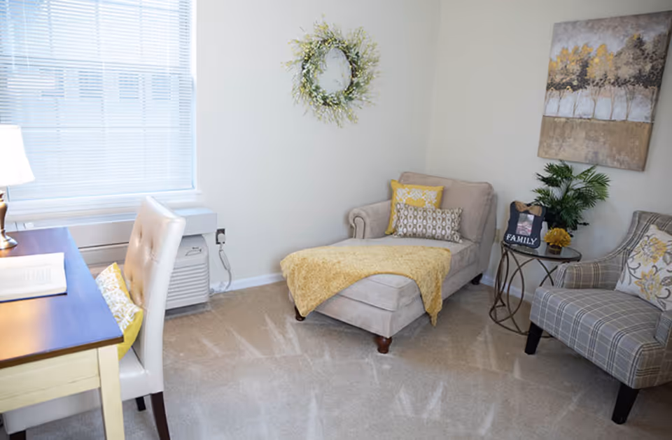 A cozy living area with a beige chaise lounge covered with a yellow throw blanket and decorative pillows, a plaid armchair with a floral pillow, a small round side table with a plant and a 'FAMILY' photo frame, a desk with a lamp and chair near a window with blinds, and wall decorations including a wreath and a painting of trees.