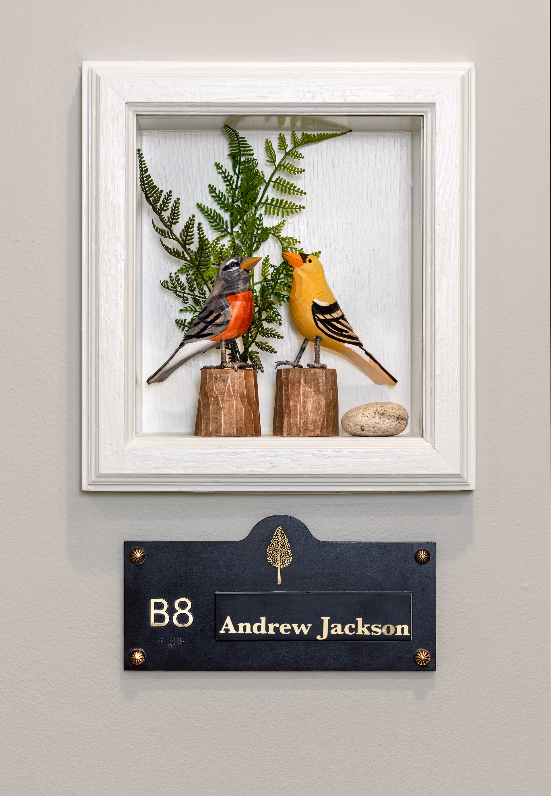 Framed display of two decorative bird figurines above a black nameplate reading "B8 Andrew Jackson" mounted on a beige wall.