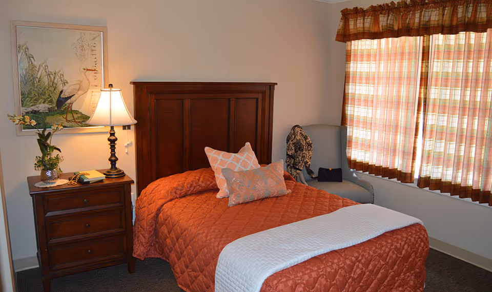 A cozy bedroom with a wooden headboard, orange quilted bed, nightstand with a lamp and plant, and a window with plaid curtains.