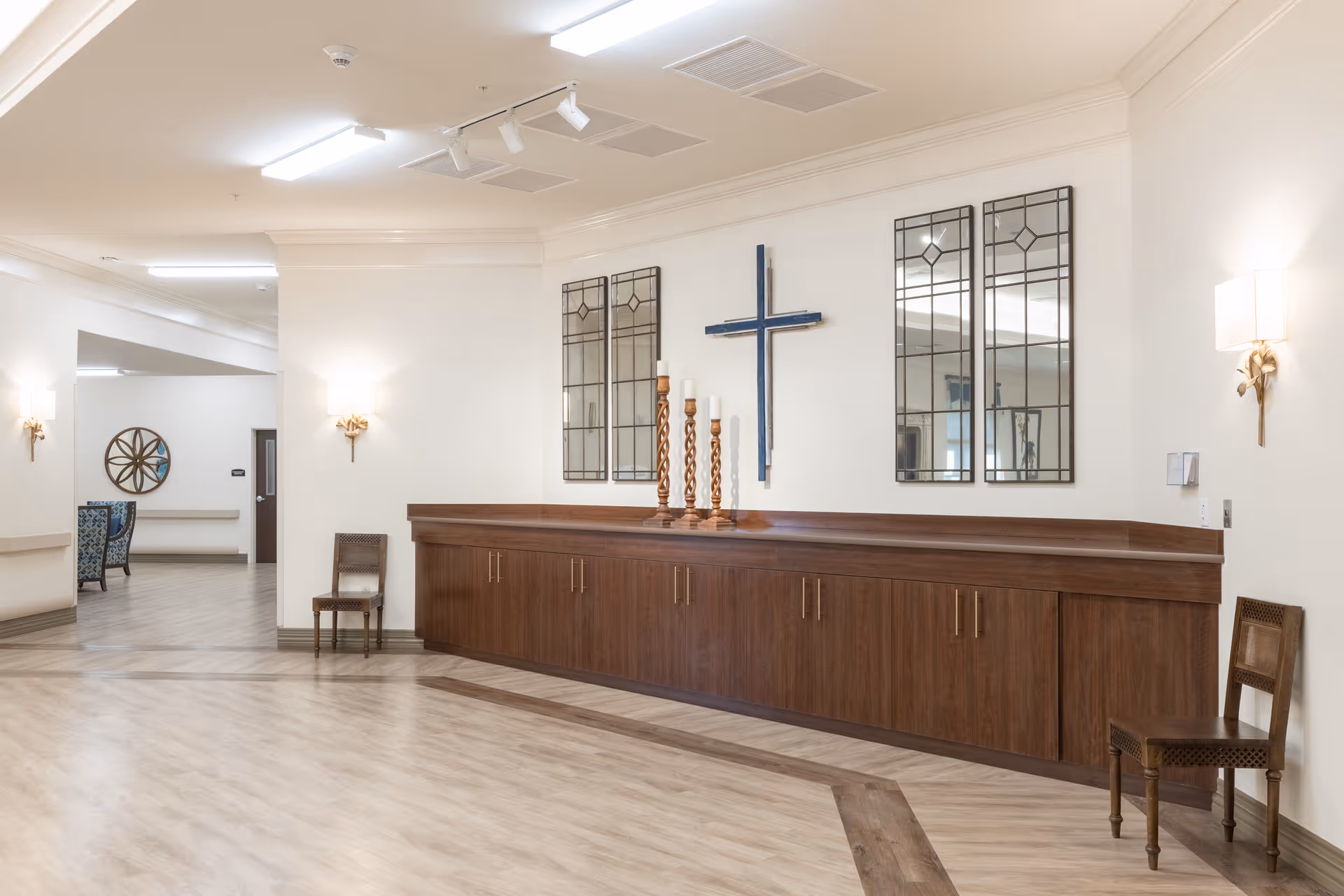 Bright interior common area with a long wooden cabinet, decorative mirrors, a wall-mounted cross, chairs, and wood-look flooring.