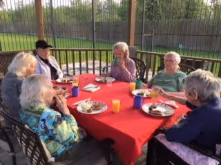 A group of six elderly people sitting around a red tablecloth-covered table outdoors, enjoying a meal together. The table has plates with food, cups, and a glass of orange juice. They are seated on metal chairs under a covered patio with a fenced background and green grass visible beyond.
