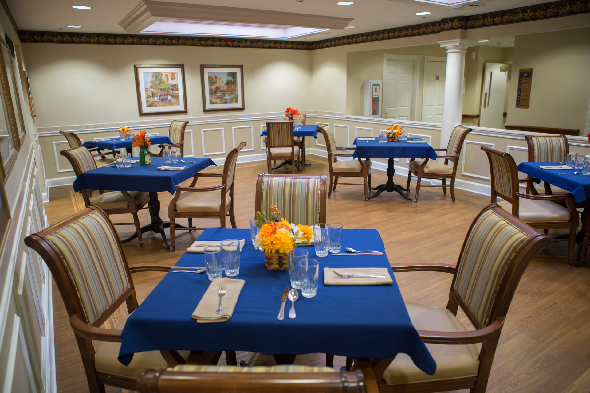 A dining room with multiple tables covered in blue tablecloths, striped chairs, and floral centerpieces.