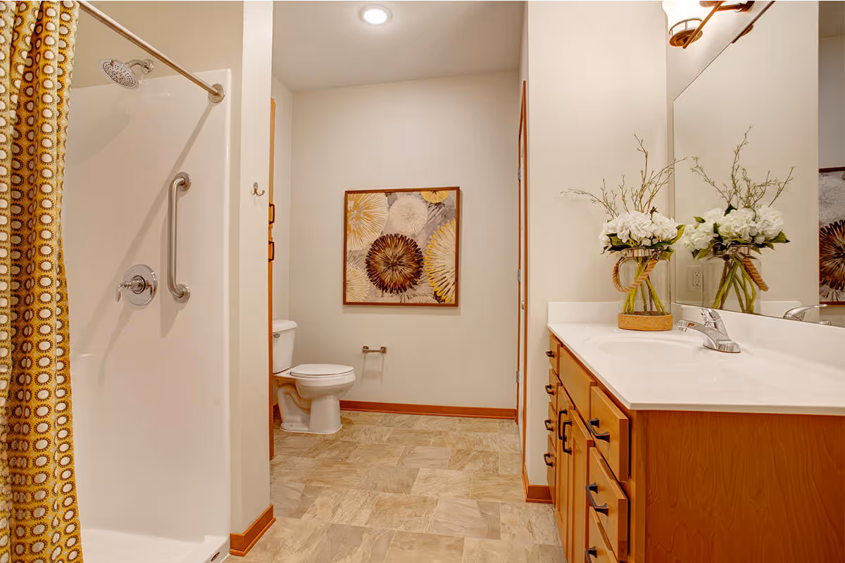 A clean and well-lit bathroom featuring a shower with a yellow patterned curtain and a grab bar, a toilet, a large mirror above a wooden vanity with a white countertop, and a vase with white flowers. A decorative framed artwork hangs on the wall opposite the vanity.