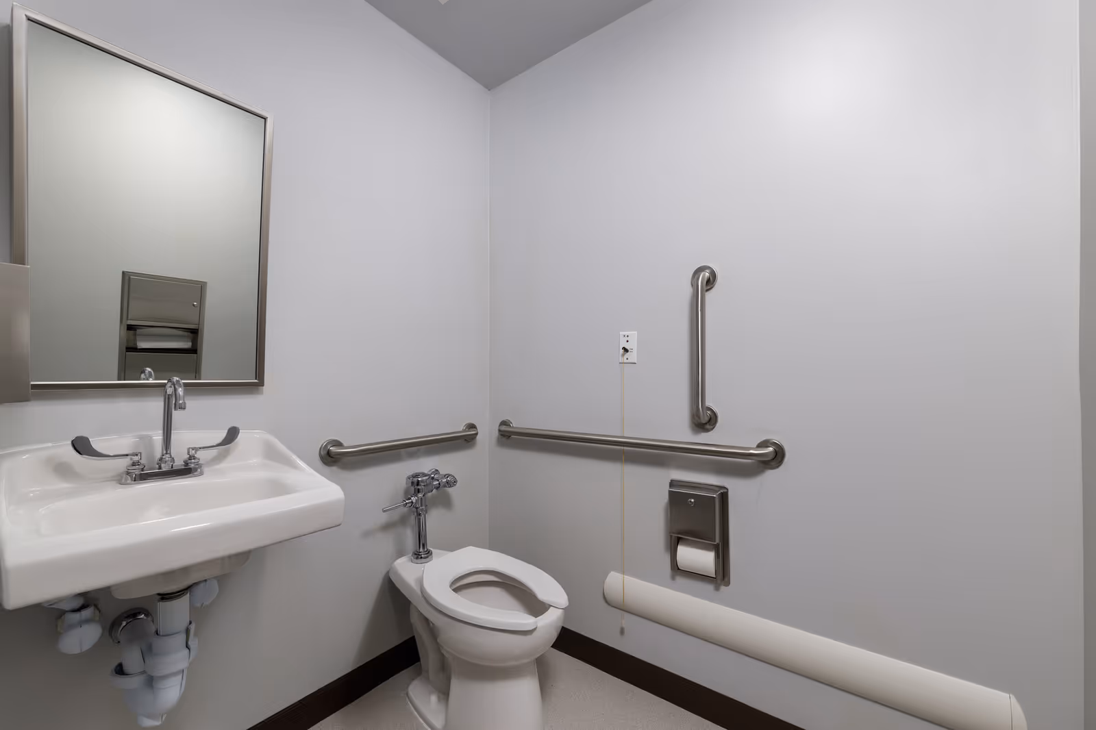 A clean, accessible bathroom with a white toilet, white sink, large mirror, and stainless steel grab bars on the walls for support. The walls are plain and light-colored, and there is a toilet paper holder mounted on the wall.
