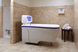 A clean, tiled bathroom featuring a modern white bathtub with a control panel, a small wooden table with neatly rolled white towels, and a separate shower area with a built-in seat.
