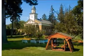 A peaceful outdoor garden area with green grass, trees, and a wooden swing set in the foreground. In the background, there is a white building with a steeple, resembling a chapel or community building, under a clear blue sky.