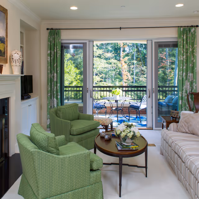 Bright living room with green upholstered chairs, a round coffee table and striped sofa facing sliding glass doors that open to a balcony with outdoor seating and a wooded view.