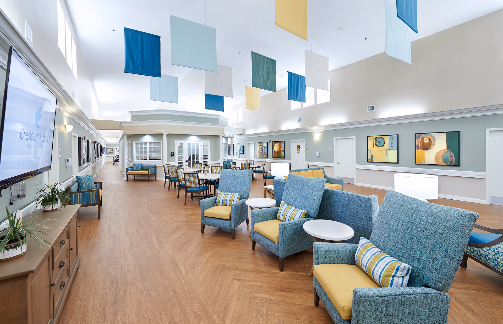 A spacious and brightly lit common area in Crescent Wood facility featuring comfortable blue and yellow upholstered chairs with small white tables, several dining tables with chairs, colorful hanging fabric panels from the ceiling, abstract wall art, and a large TV screen displaying the Crescent Wood logo.