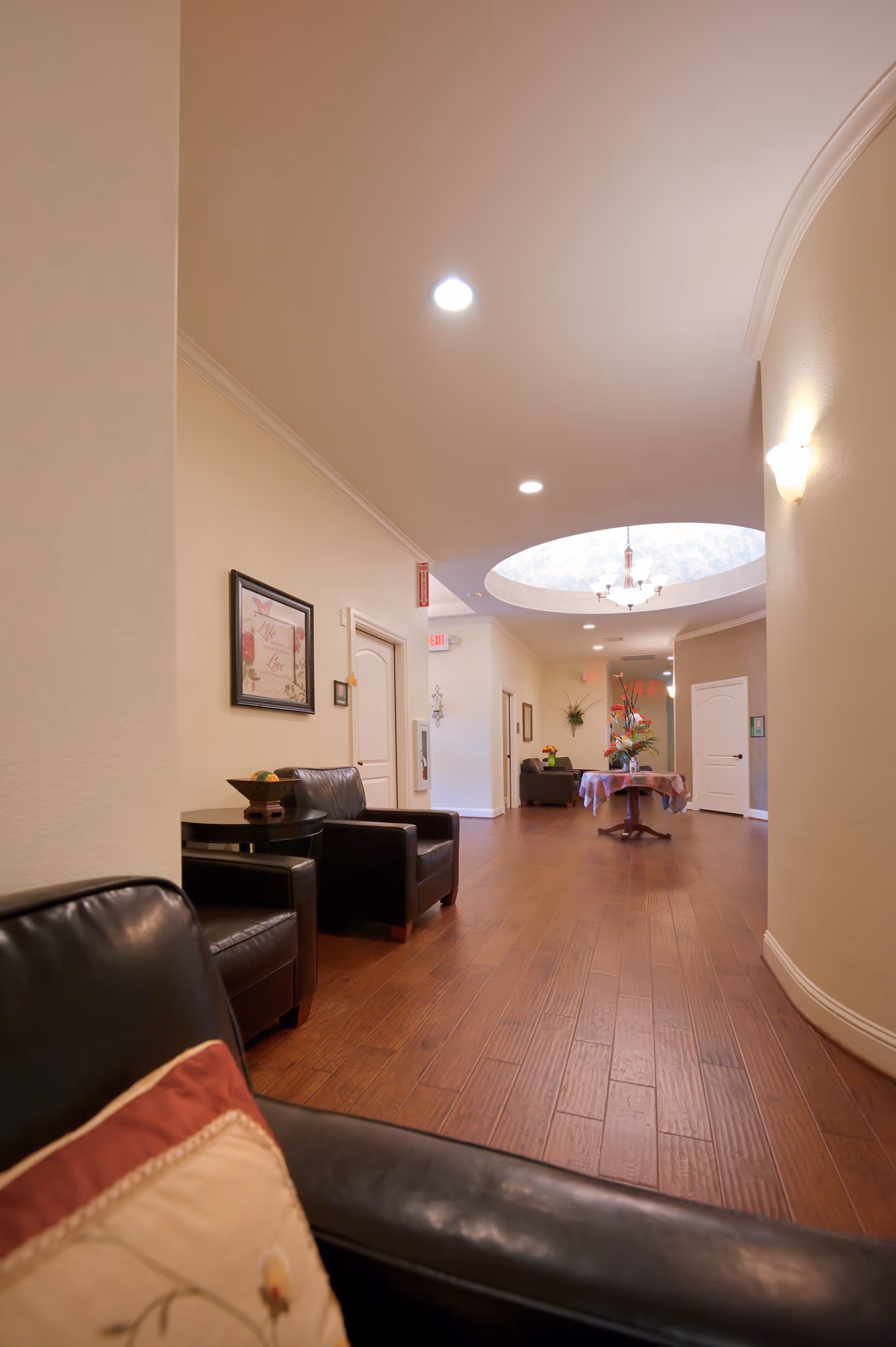 Interior hallway of a senior living facility with wooden flooring, beige walls, and recessed ceiling lights. There are black leather chairs with a small table and decorative items along the wall. A round table with a floral arrangement is placed under a circular skylight with a chandelier. Several doors and wall decorations are visible along the hallway.
