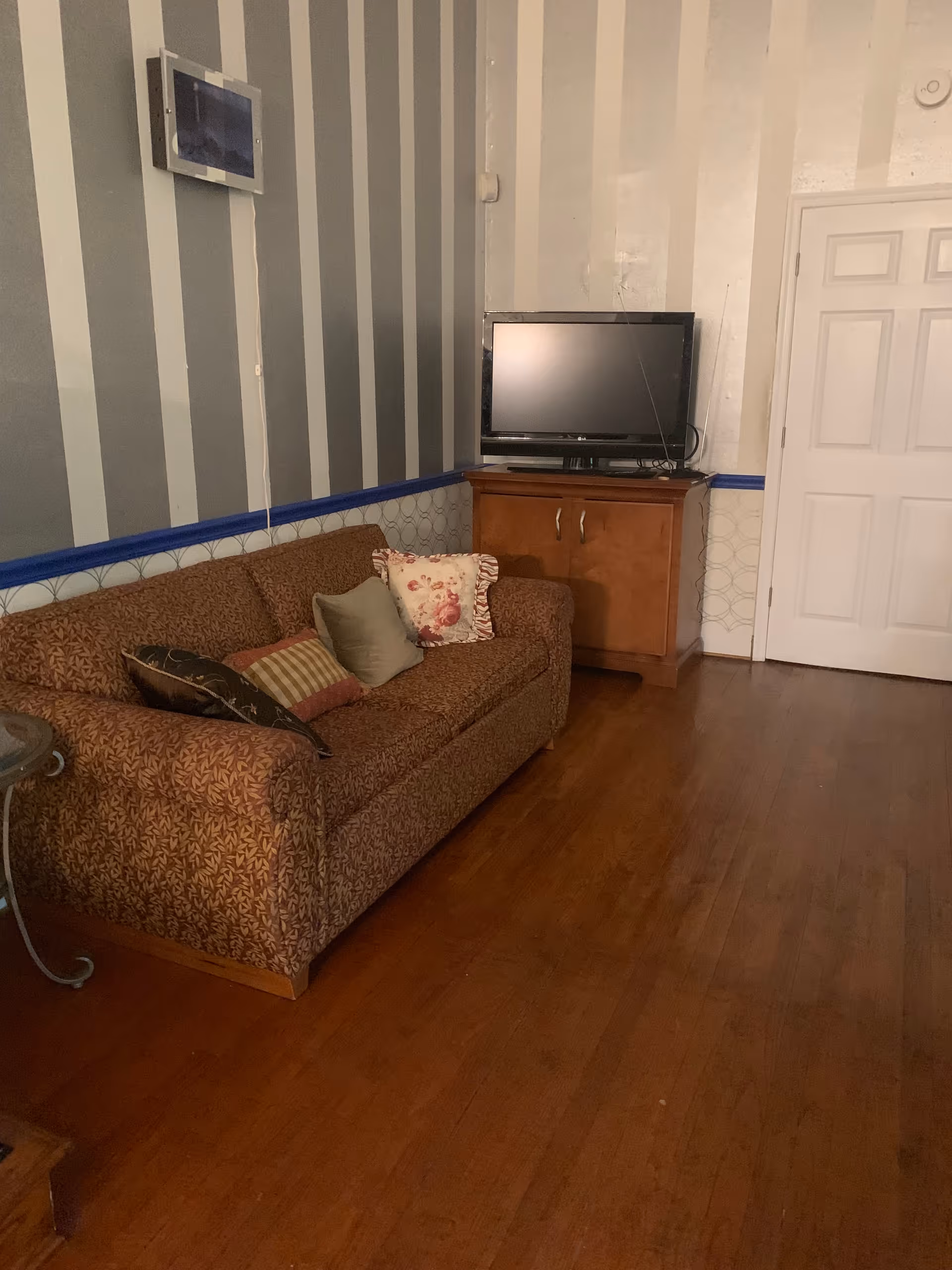 A living room with a patterned brown sofa adorned with several cushions, a wooden cabinet with a flat-screen TV on top, striped wallpaper in shades of gray and white, and a wooden floor. There is a white door on the right side of the room.