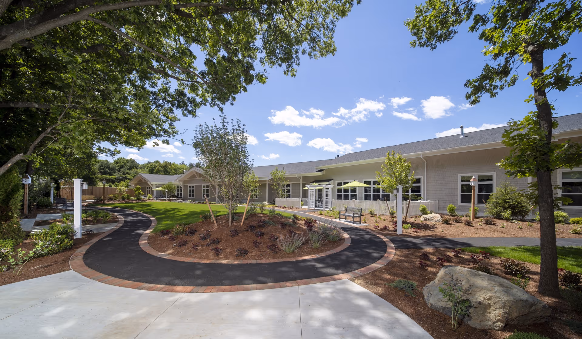 Outdoor view of a senior living facility with a curved paved walkway surrounded by landscaped gardens, trees, and benches. The building is single-story with large windows and a light-colored exterior under a blue sky with scattered clouds.