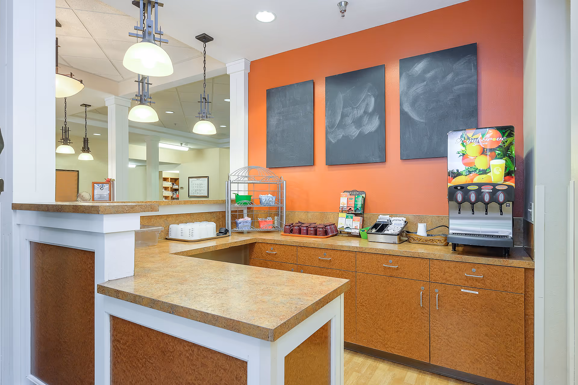 A beverage and snack counter area with a juice dispenser, a toaster, cups, and various condiments on a countertop with wooden cabinets below. The wall behind the counter is painted orange and has three blank chalkboards hanging on it. Pendant lights hang from the ceiling, and a mirror reflects part of the room.